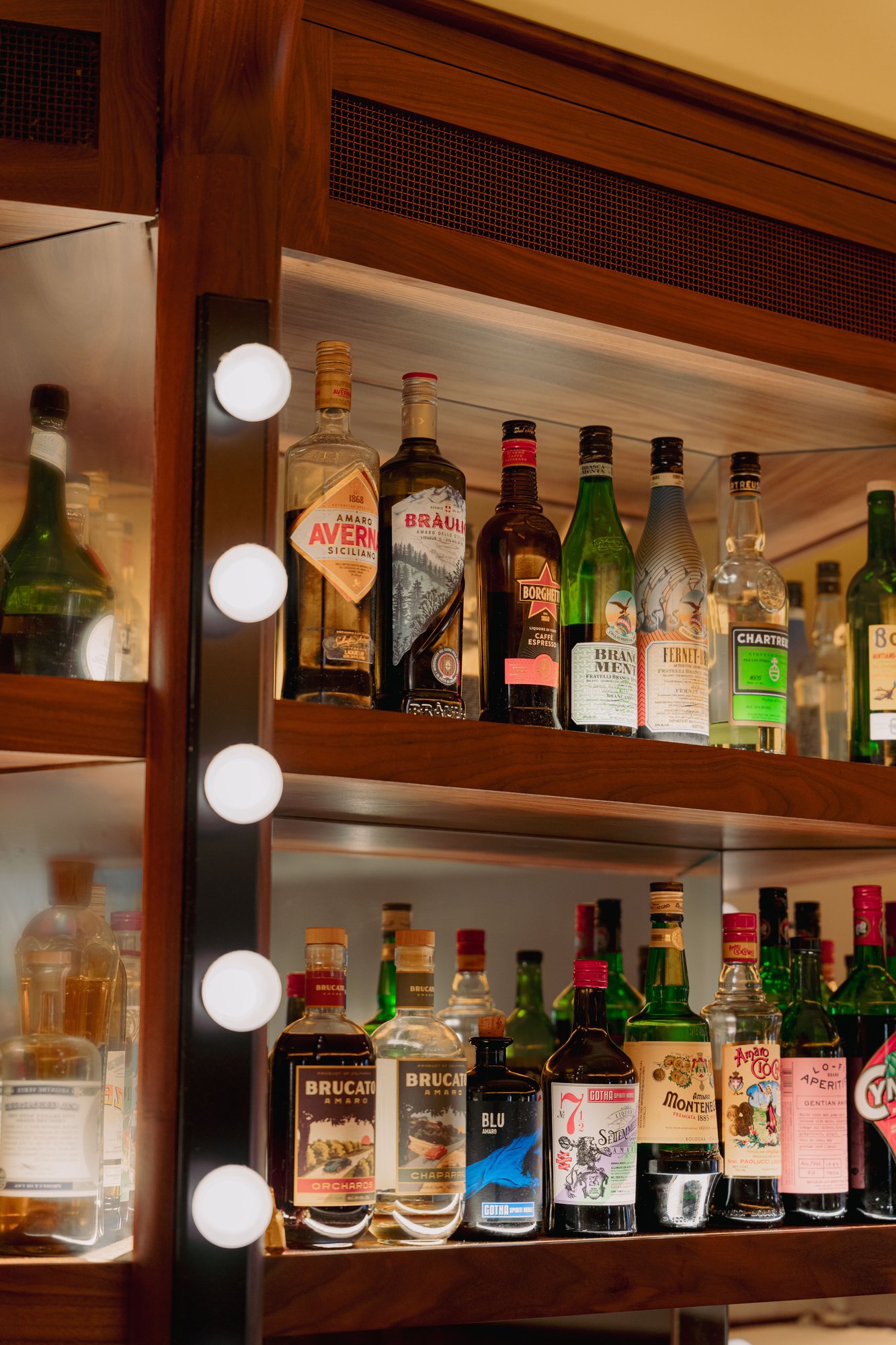 Rich back bar shelving with round vanity bulb lighting displaying amaro bottles including Averna, Braulio, Fernet Branca, and Chartreuse
